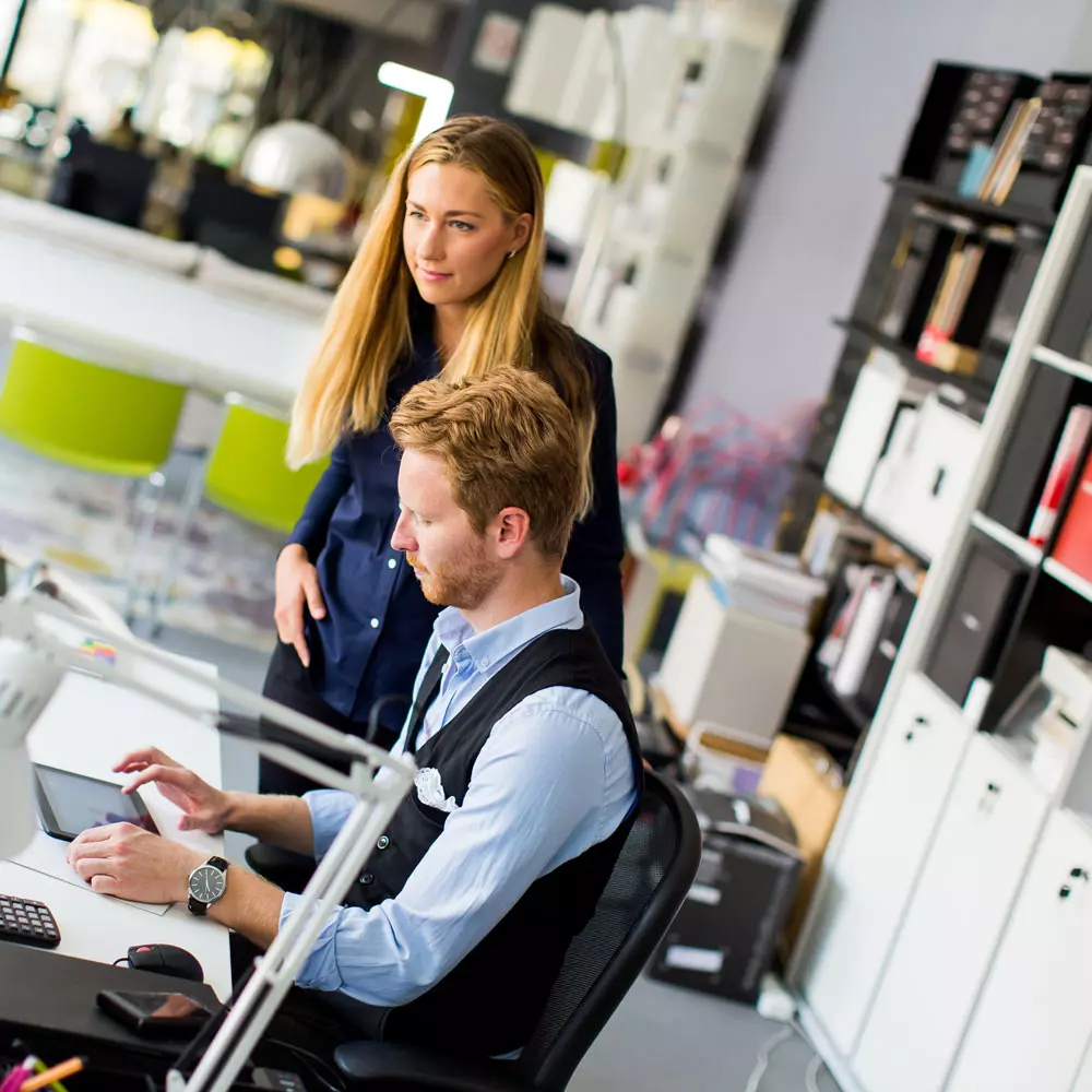 Lees meer Een collega staat naast een bureau waarachter een andere collega zit. Ze kijken beiden naar een tablet.