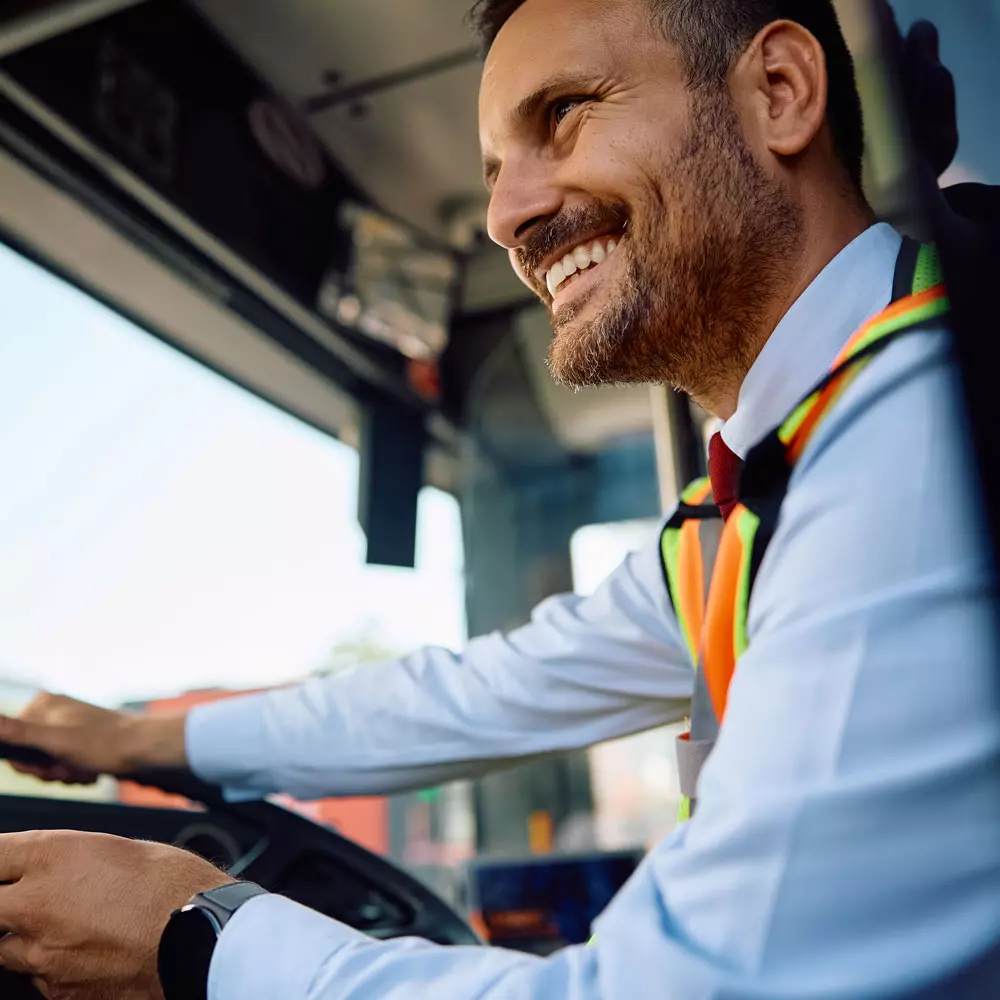 Lees meer Een chauffeur in een nette shirt, stropdas en oranje veiligheids hesje lacht terwijl hij een vrachtwagen bestuurt.
